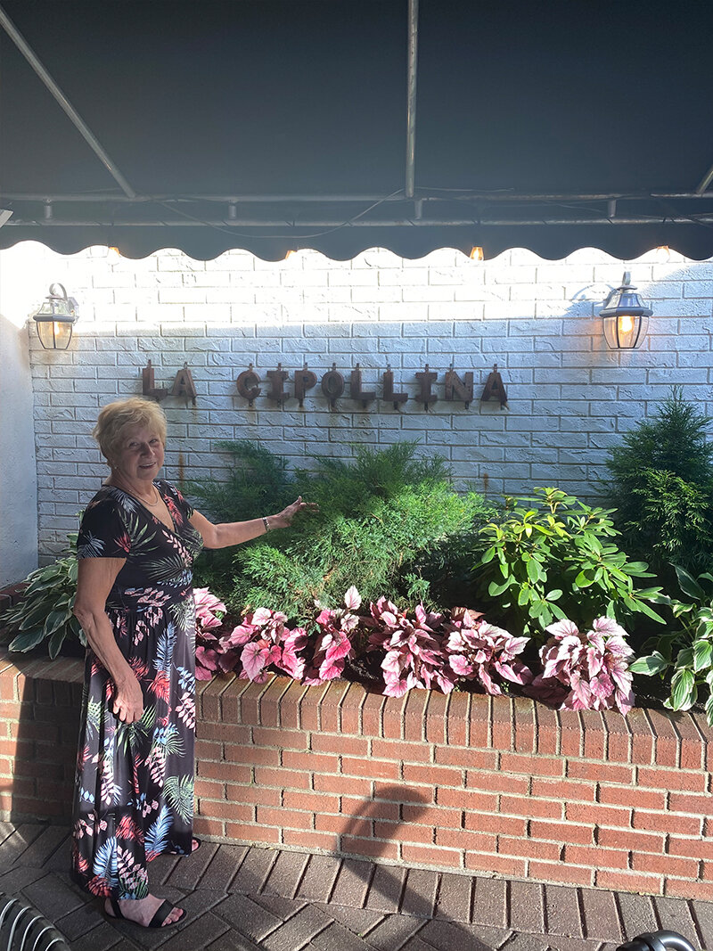 Woman standing in front of bushes pointing to La Cipollina sign